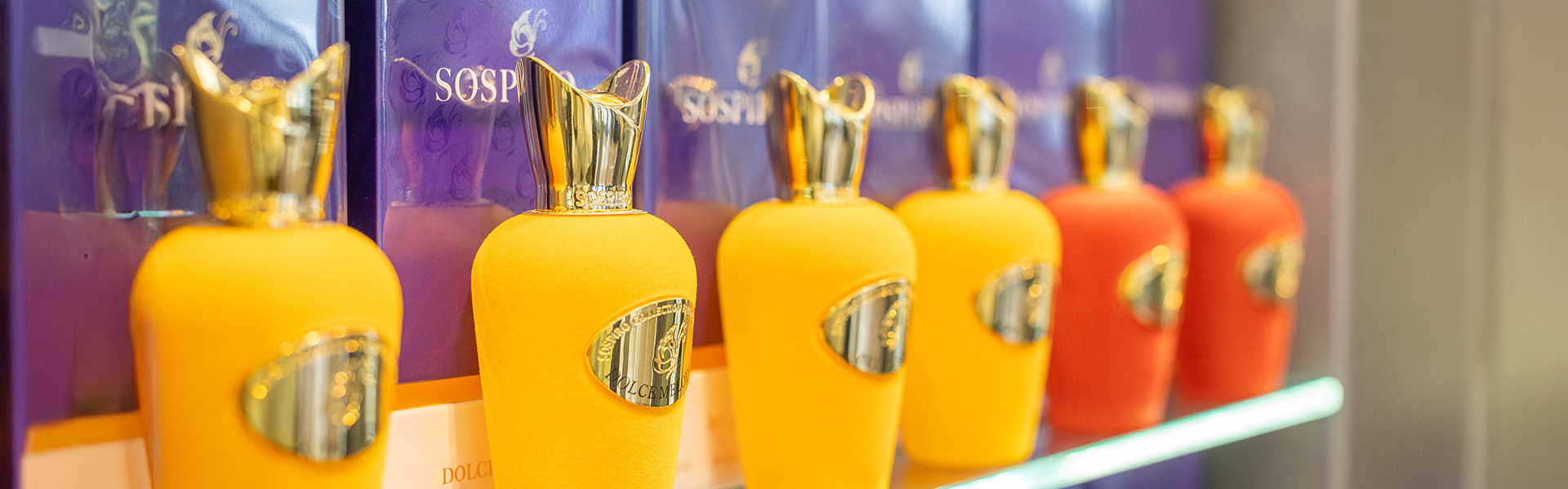Sospiro - Reihe bunter Parfumflaschen mit goldenen Verschlüssen, vor lila Verpackungsschachteln ausgestellt.